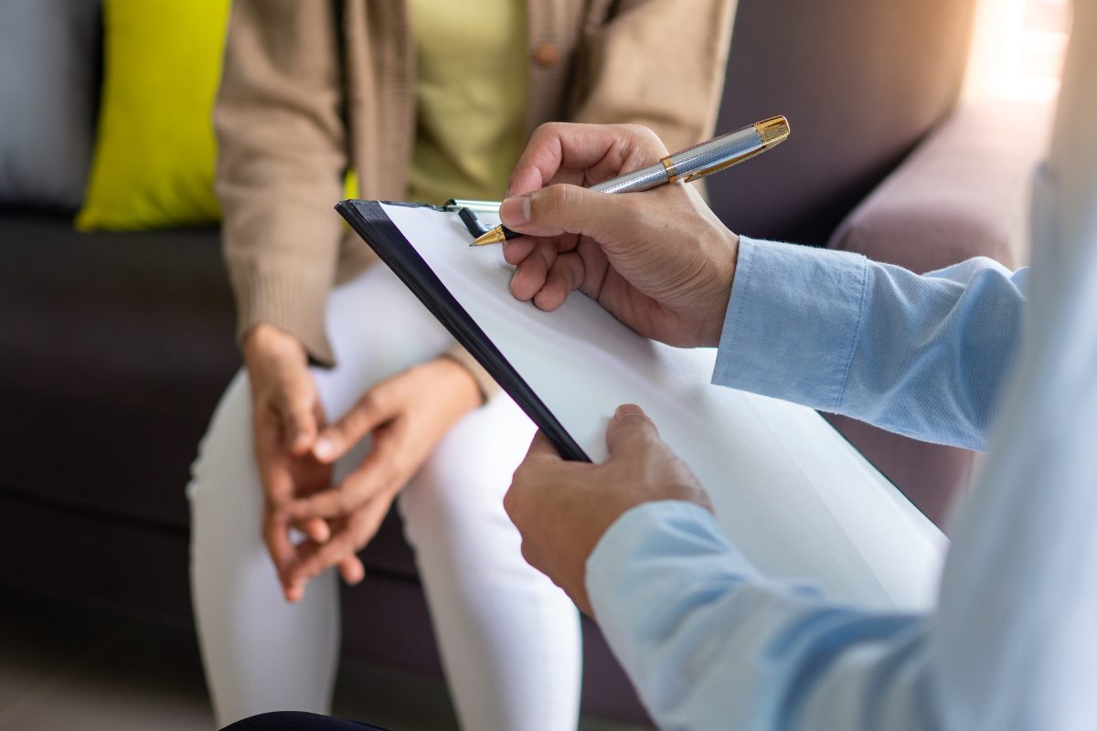 Clinician writing notes during a mental health evaluation with a client seated nearby, illustrating the documentation process for a mental status exam.