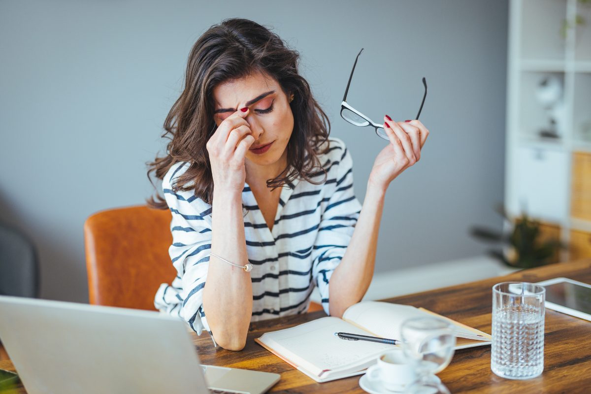 Exhausted therapist experiencing burnout and compassion fatigue, holding glasses while rubbing bridge of nose at desk, illustrating the emotional toll of clinical work.