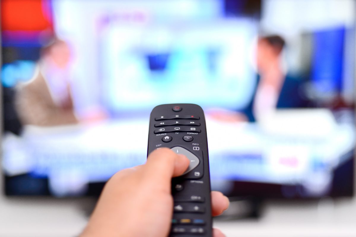 Hand holding a TV remote pointed at political news broadcast, symbolizing political obsession disorder